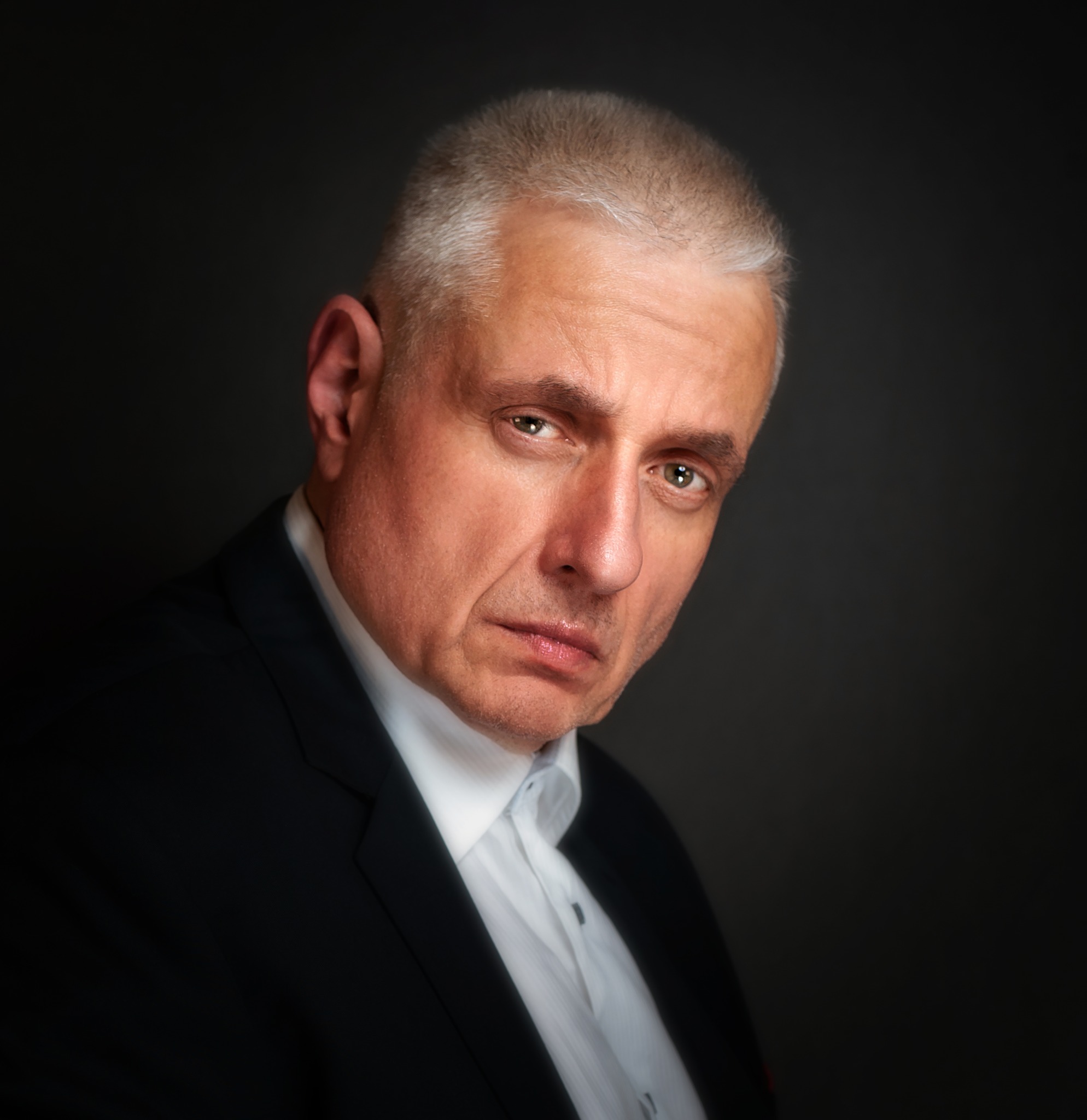 Portret mężczyzny w garniturze i białej koszuli, z krótkimi siwymi włosami, na ciemnoszarym tle.