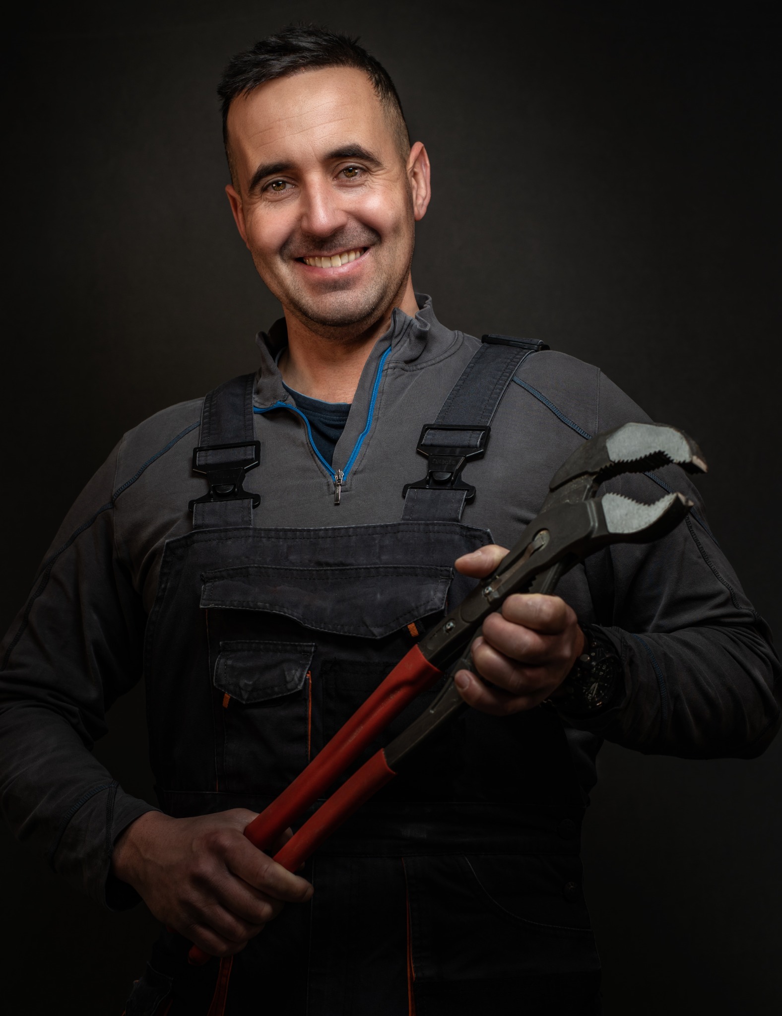 Uśmiechnięty mężczyzna w kombinezonie roboczym trzymający duże, regulowane szczypce na ciemnym tle.