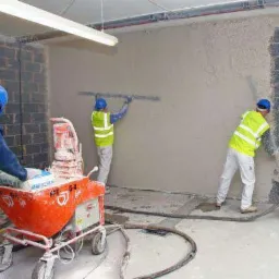 Ekipa budowlana w trakcie nakładania tynku maszynowego na ścianę, widoczna pomarańczowa maszyna do tynkowania, jeden pracownik wyrównuje tynk łatą, drugi rozprowadza tynk pistoletem