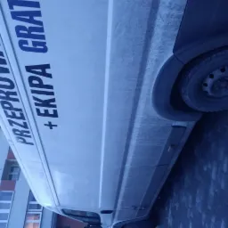 Biały samochód dostawczy z niebieskim napisem 'PRZEPROWADZKI + EKIPA GRAT' zaparkowany na brukowanej ulicy, z budynkiem w tle.