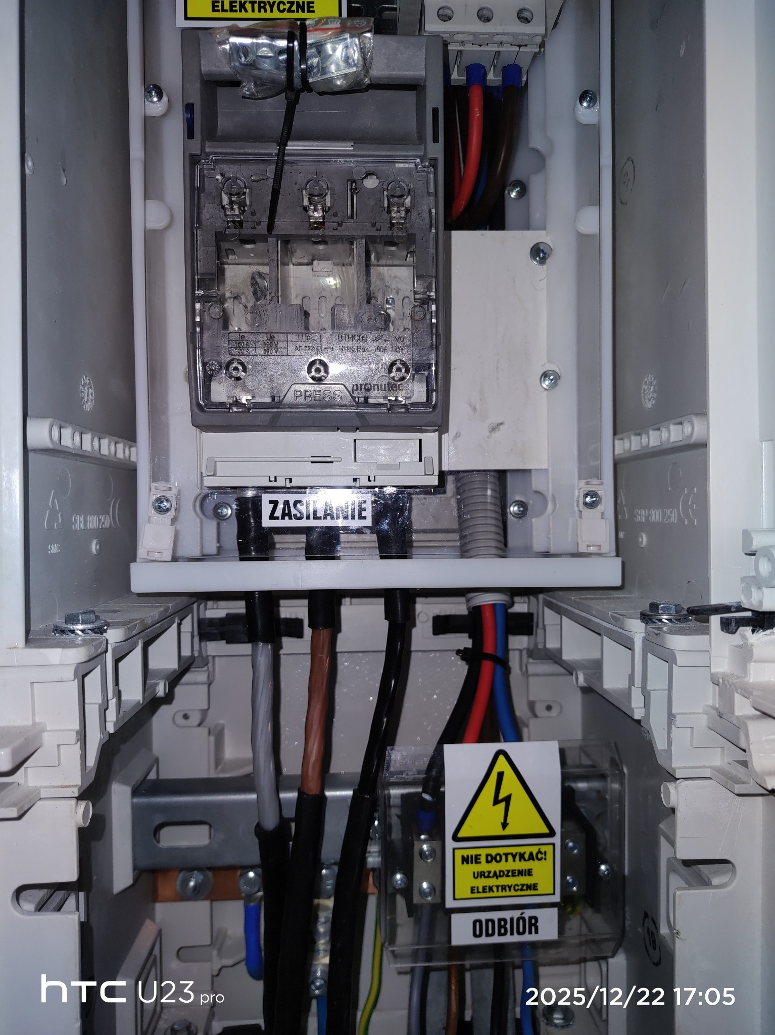 Wnętrze skrzynki elektrycznej z przewodami, bezpiecznikami i oznaczeniami 'Zasilanie' i 'Odbiór', widoczne kable w kolorach czerwonym, niebieskim i brązowym.