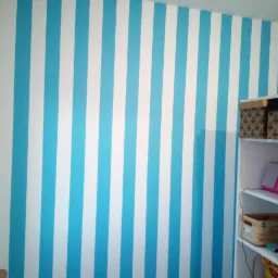 Ściana w pokoju dziecięcym z równymi, pionowymi pasami w kolorze błękitnym i białym, obok biały regał z pudełkami i różowym radiem.