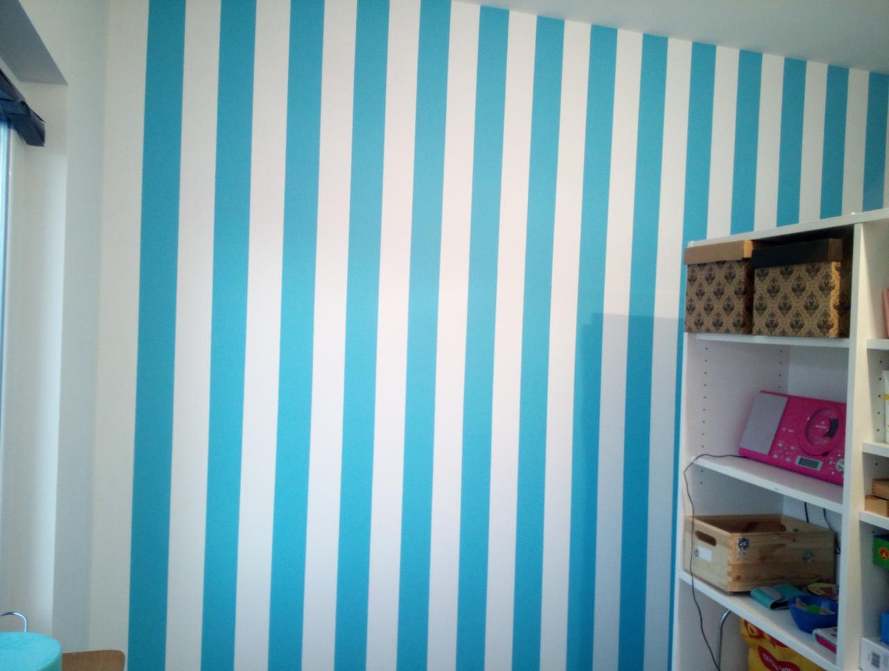 Ściana w pokoju dziecięcym z równymi, pionowymi pasami w kolorze błękitnym i białym, obok biały regał z pudełkami i różowym radiem.