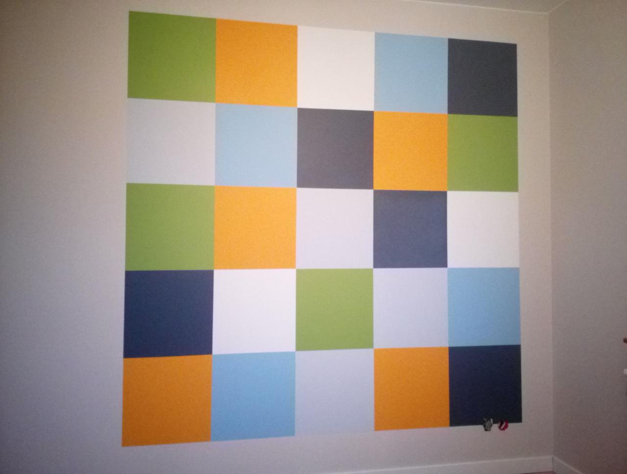 Ściana z kwadratami w różnych kolorach: zielonym, pomarańczowym, białym, niebieskim i szarym, tworzącymi geometryczny wzór w pokoju.