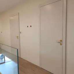 Dwa białe, minimalistyczne drzwi wewnętrzne z złotymi klamkami, widoczne z perspektywy korytarza z drewnianą podłogą i szklaną balustradą. Otwory na gniazdka.