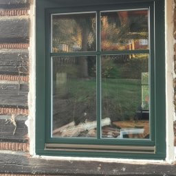 Zakład Stolarski Andrzej Górski - Zamontowane okno z zieloną ramą w drewnianej ścianie domu. Widoczne uszczelnienie pianką montażową. W szybach odbija się zieleń ogrodu.