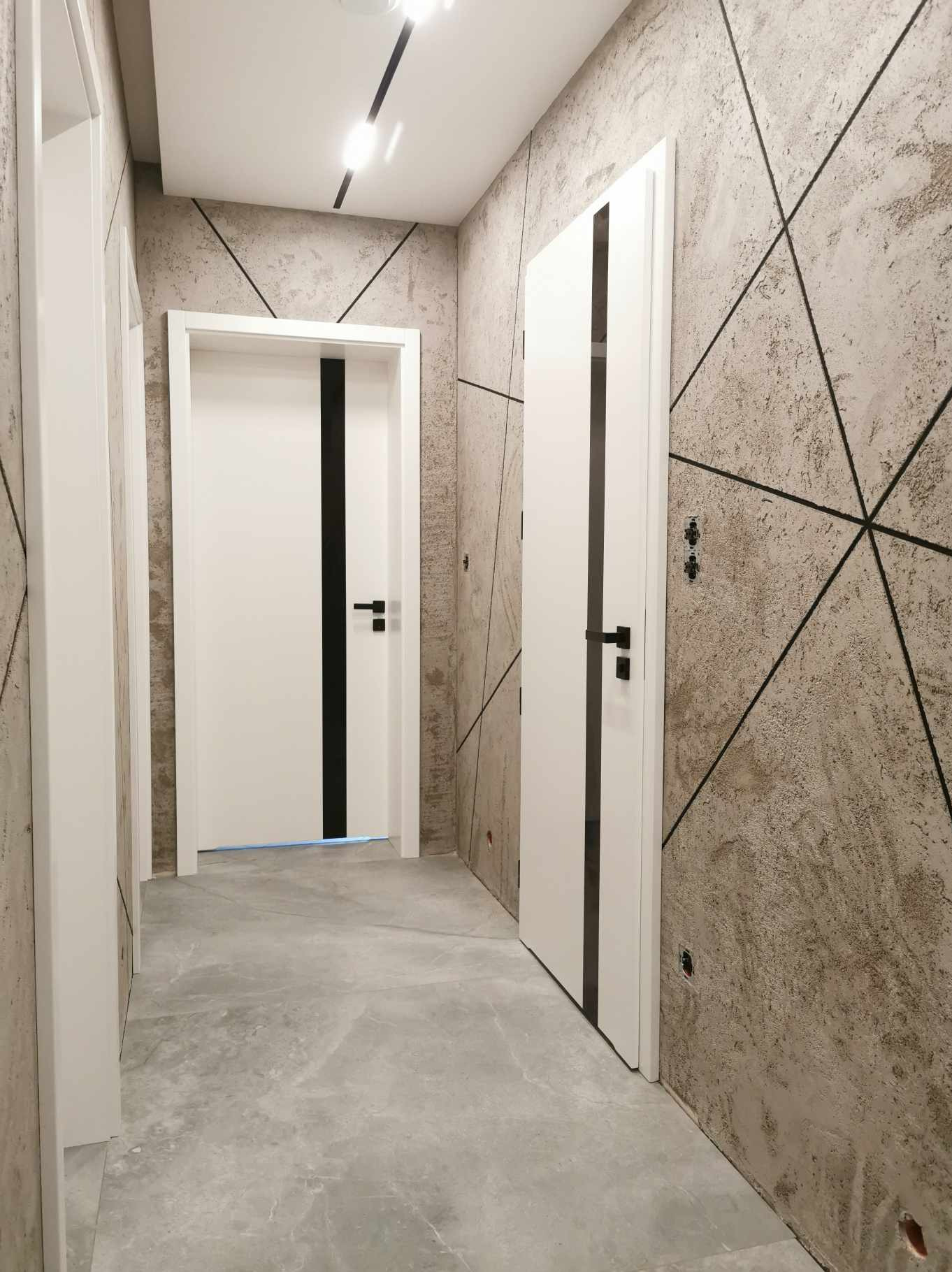 Nowoczesne, białe drzwi wewnętrzne z czarnym pionowym pasem, minimalistyczna klamka, na tle ściany z dekoracyjnym wzorem geometrycznym i szarą podłogą z efektem kamienia.