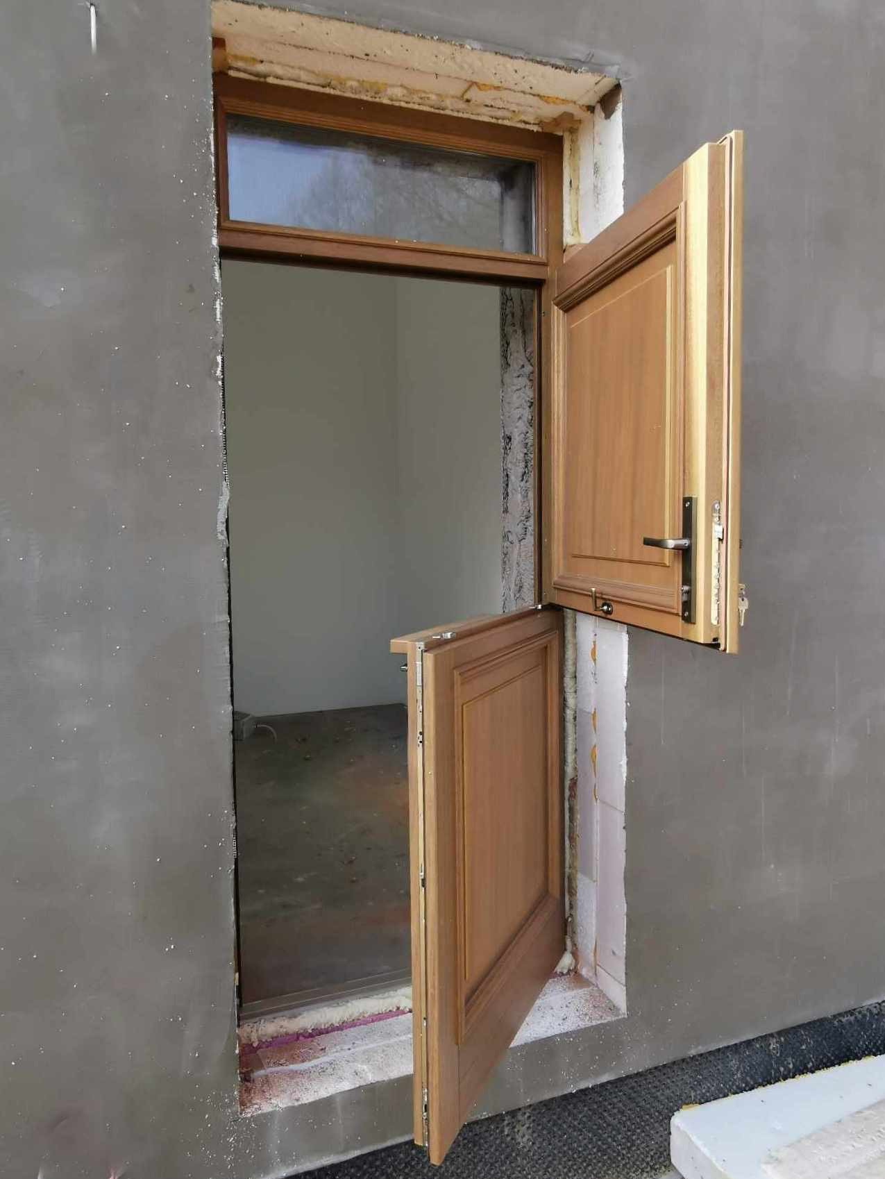 Montaż dwuczęściowych drzwi drewnianych z oknem nad drzwiami w surowej ścianie, widoczne pianka montażowa i niedokończone wykończenie.