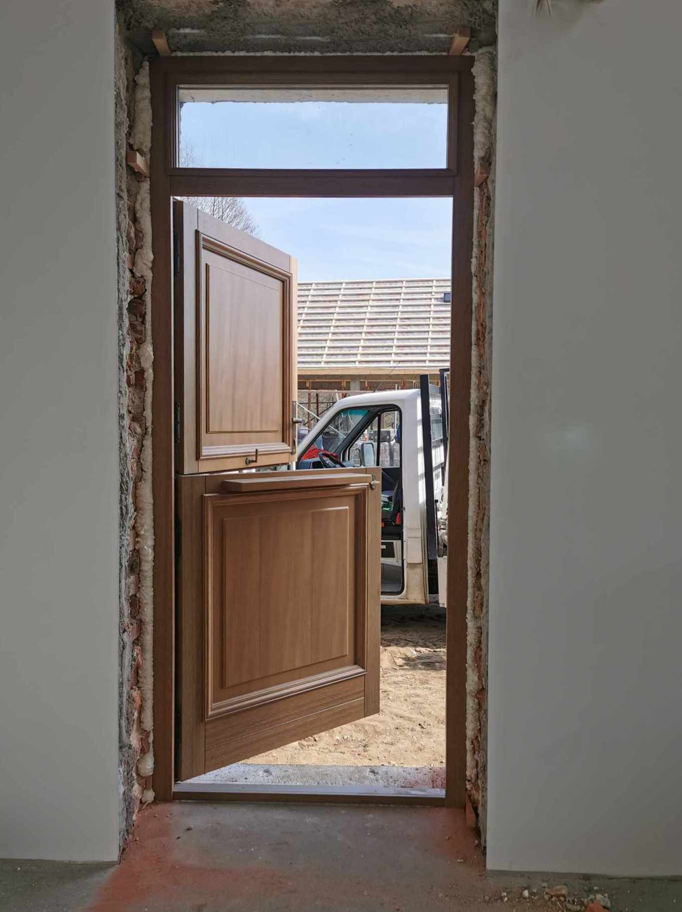 Montaż dwuczęściowych, drewnianych drzwi w surowym stanie wykończenia, widoczna piana montażowa i cegły, w tle samochód dostawczy i fragment dachu.