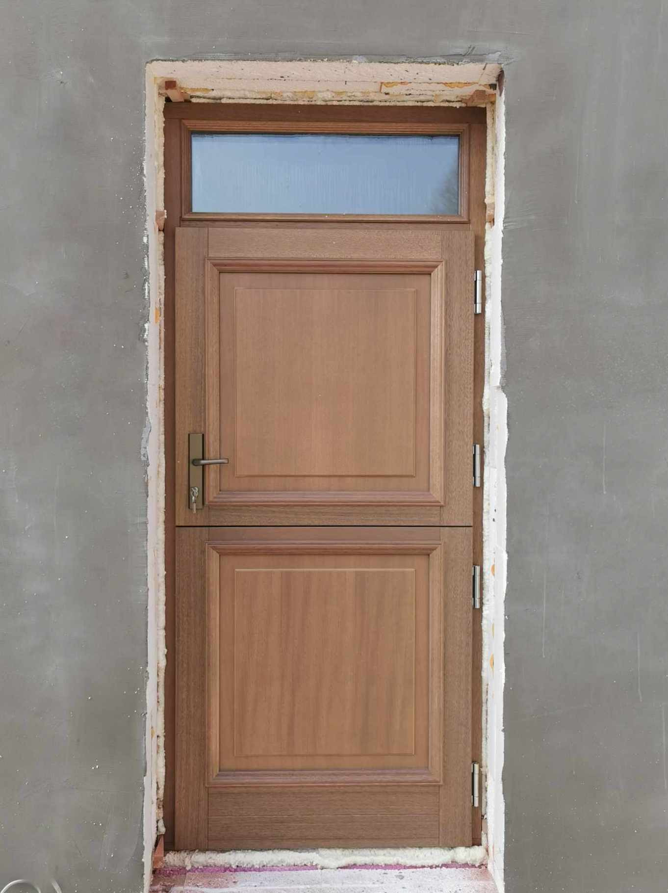 Brązowe drzwi wejściowe z górnym oknem zamontowane w surowym, szarym murze, widoczne ślady pianki montażowej i uszczelnienia.
