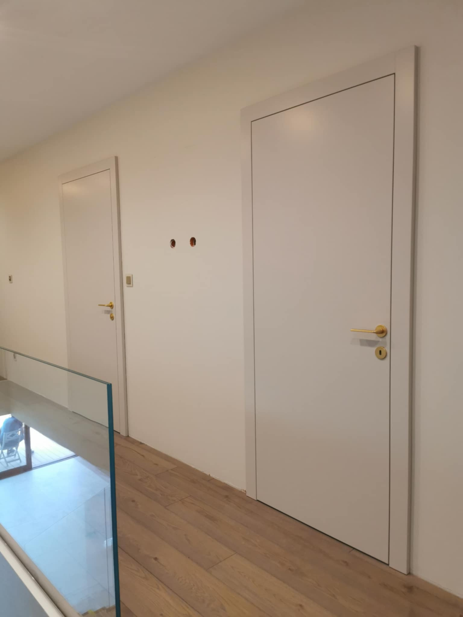 Dwa białe, minimalistyczne drzwi wewnętrzne z złotymi klamkami, widoczne z perspektywy korytarza z drewnianą podłogą i szklaną balustradą. Otwory na gniazdka.