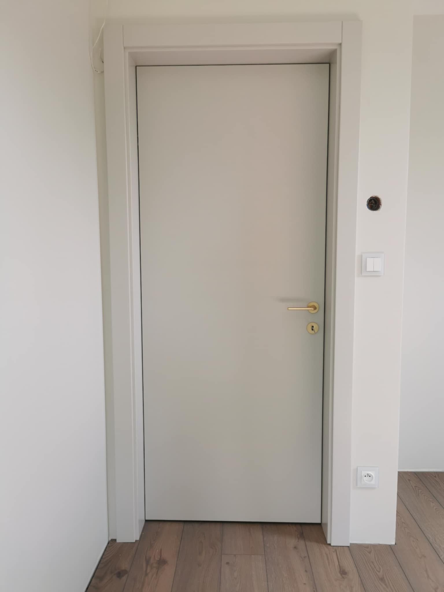Zamknięte, gładkie drzwi wewnętrzne w białej futrynie z złotą klamką na tle białej ściany i drewnianej podłogi. Widoczny kontakt i włącznik światła.