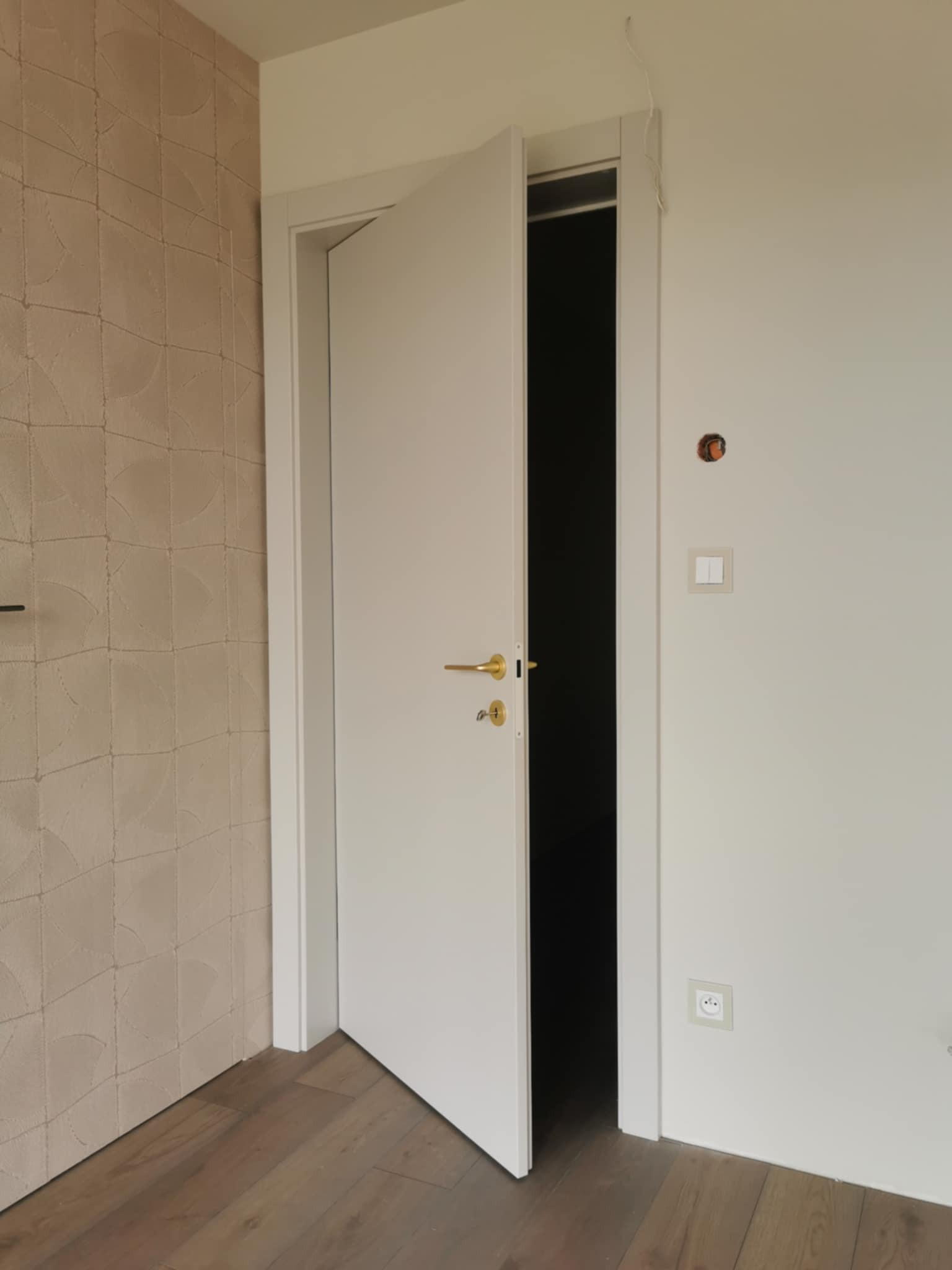 Otwarta, biała, minimalistyczna drzwi wewnętrzne z złotą klamką, widoczne na tle jasnych ścian i drewnianej podłogi. Ściana po lewej stronie z beżową fakturą.