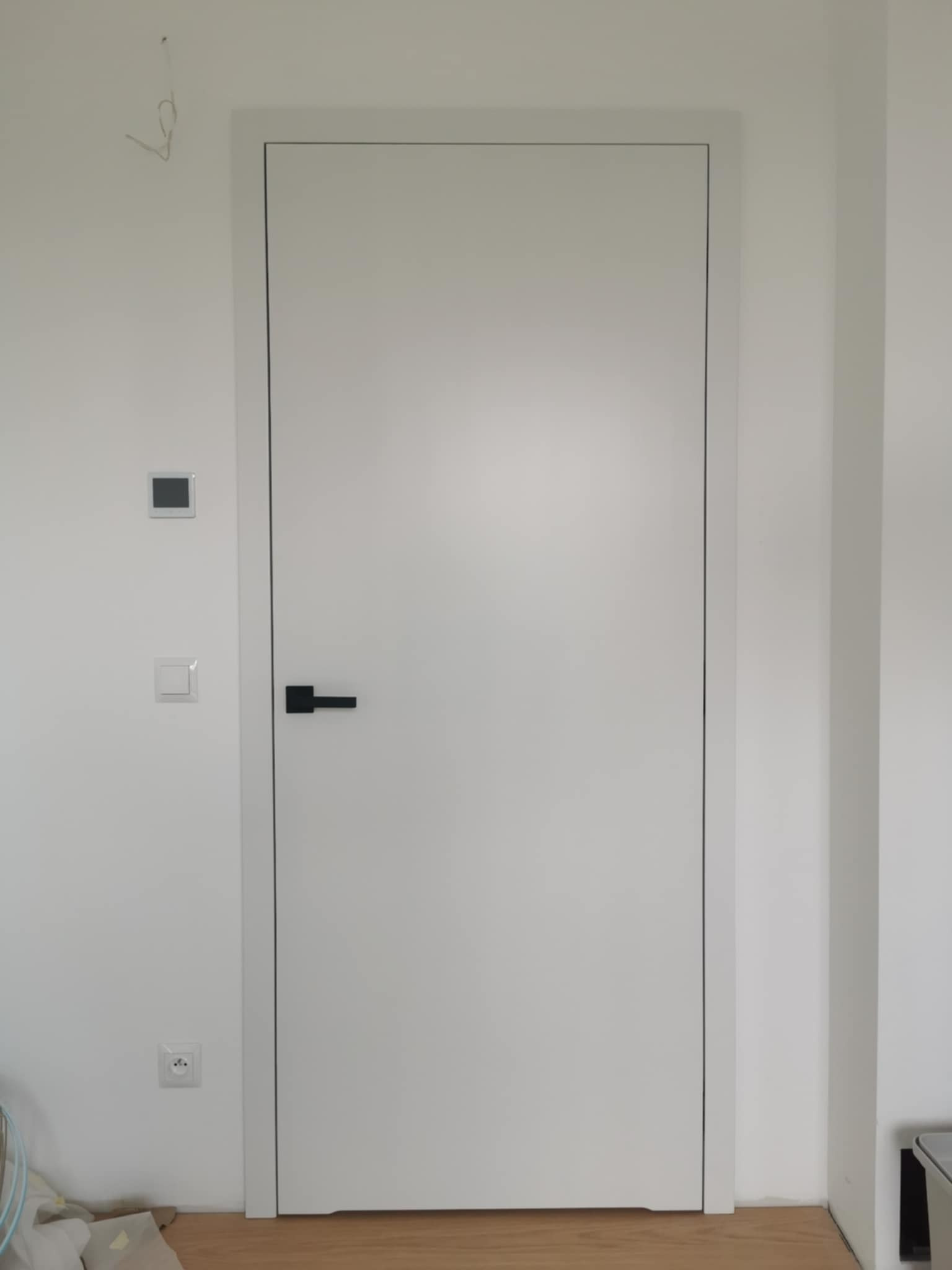 Białe drzwi wewnętrzne z czarną klamką, osadzone w białej ścianie z widocznymi włącznikami i gniazdkami, na drewnianej podłodze. Minimalistyczny design.