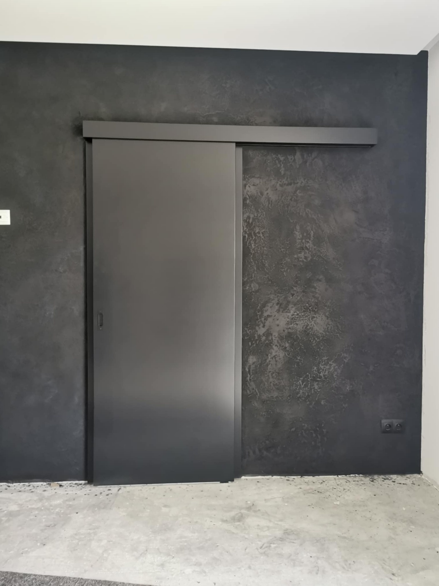 Szare, minimalistyczne drzwi przesuwne w ciemnym pomieszczeniu z betonową posadzką. Ściana z dekoracyjną fakturą. Widoczny fragment gniazdka elektrycznego.
