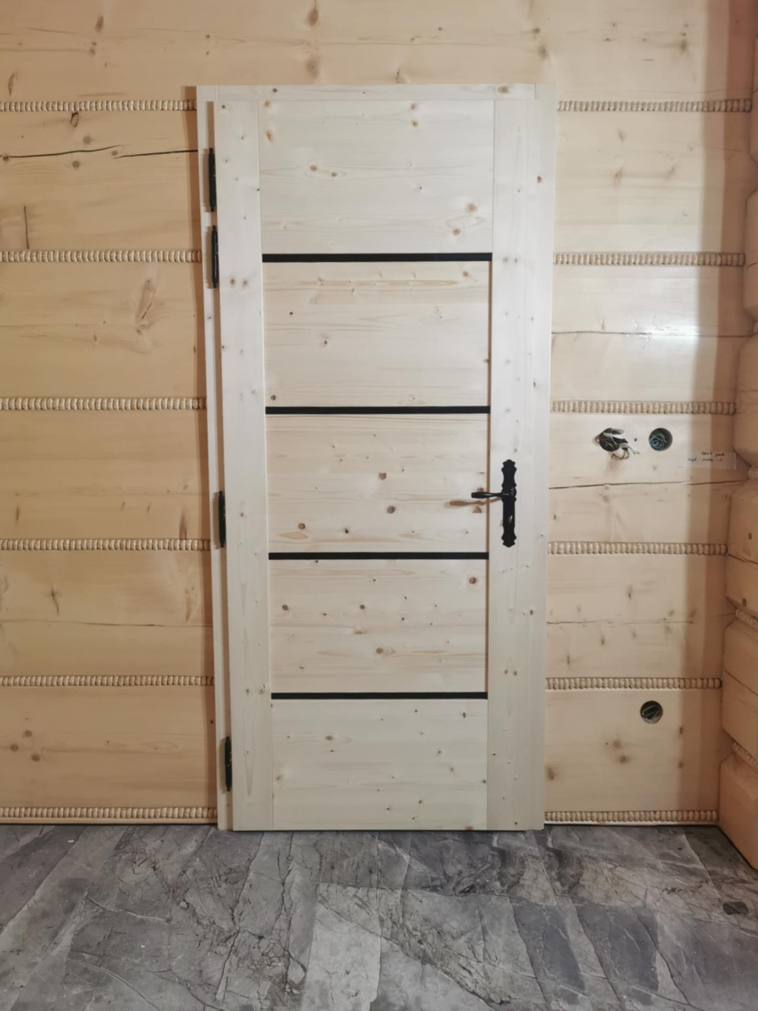 Jasne, drewniane drzwi wewnętrzne z czarnymi, poziomymi akcentami, zamontowane w ścianie z boazerii. Widoczna czarna klamka i zawiasy.