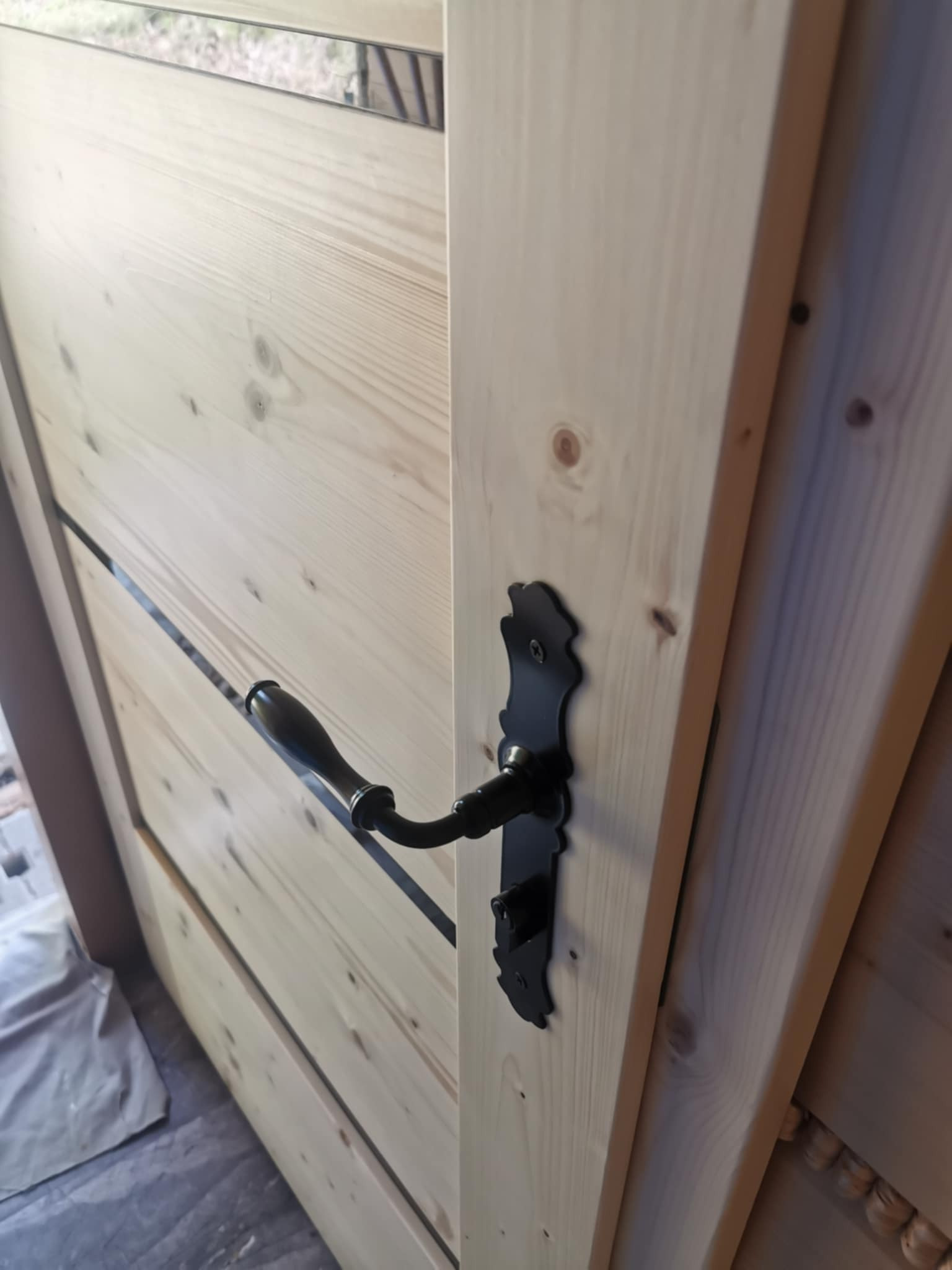 Jasne, surowe drewno drzwi wewnętrznych z czarną, ozdobną klamką. Widoczne słoje drewna i detale wykończenia. Minimalistyczny design z elementami rustykalnymi.