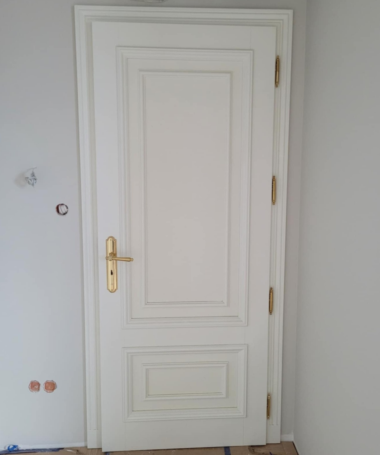 Eleganckie, białe drzwi wewnętrzne z ozdobnymi frezami i złotą klamką, zamontowane w jasnym wnętrzu. Widoczne detale wykończenia i zawiasy.