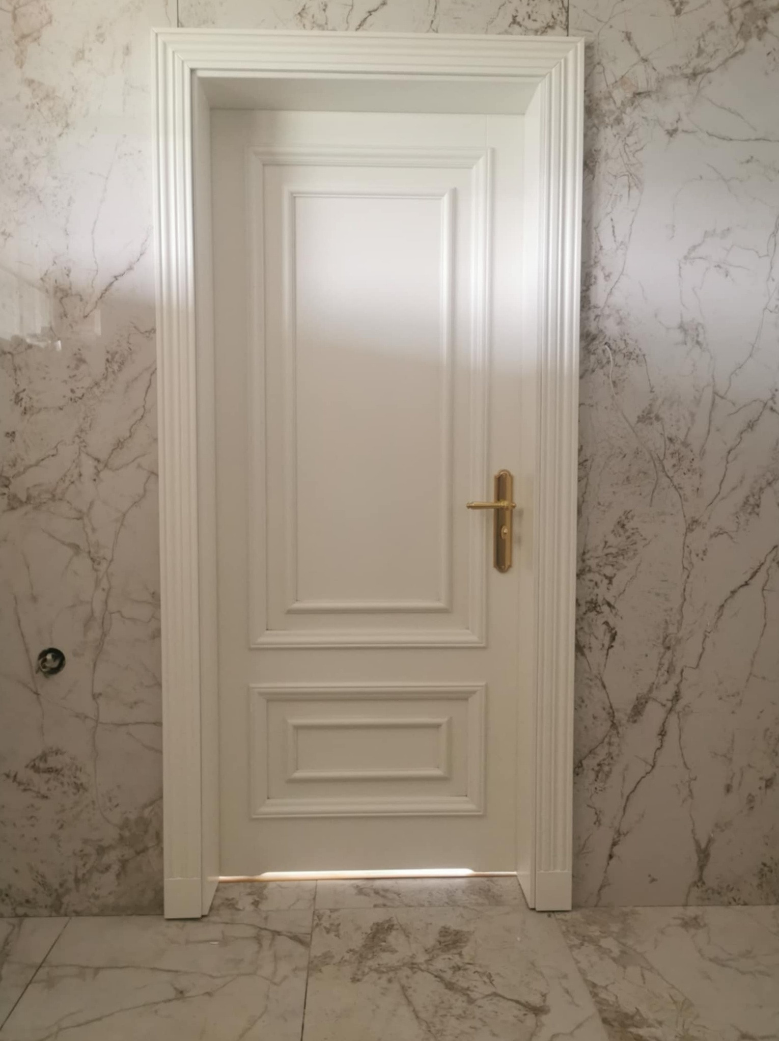 Białe, klasyczne drzwi wewnętrzne z ozdobnymi frezami i złotą klamką, zamontowane w łazience wykończonej marmurowymi płytami.