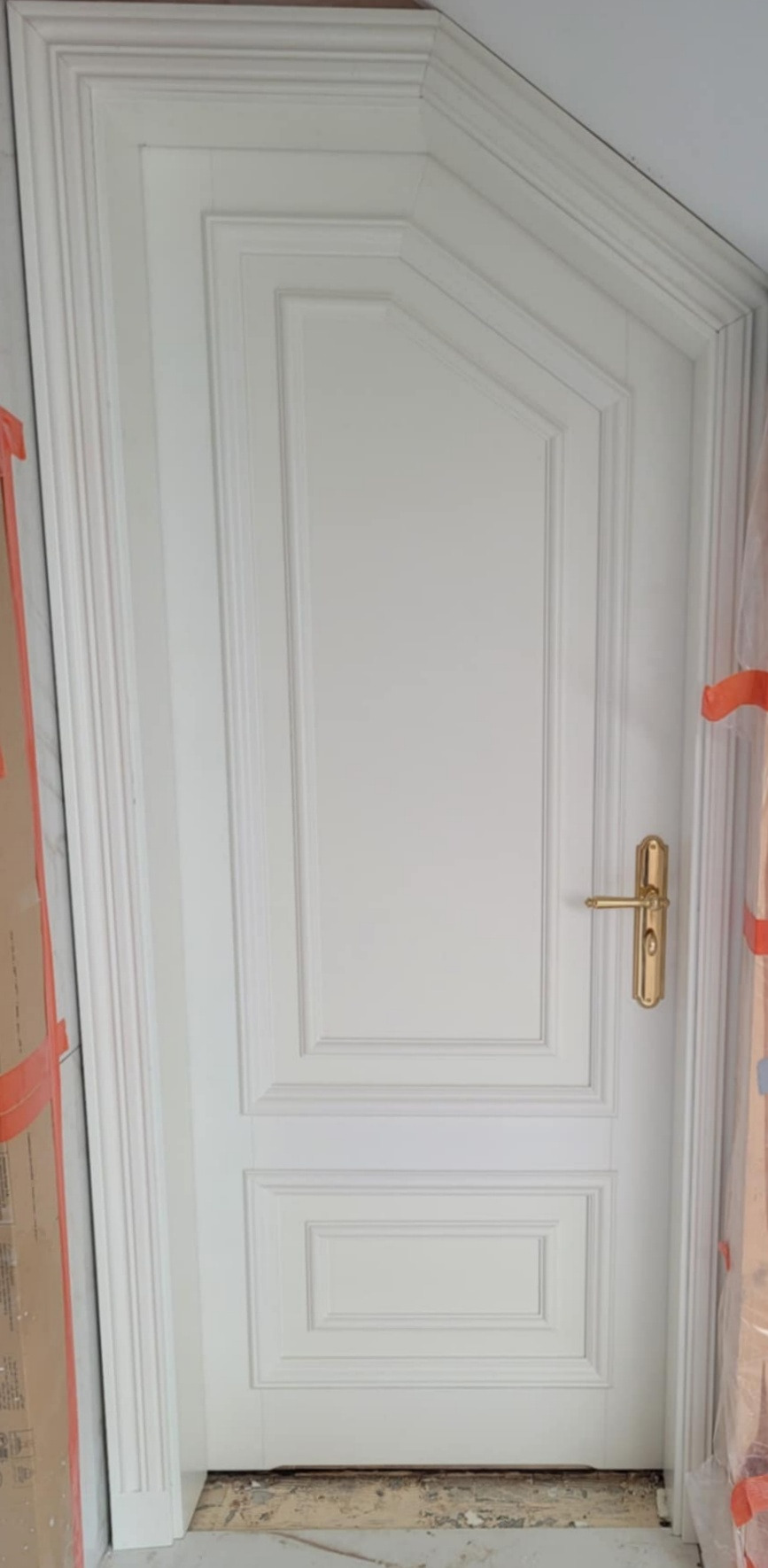 Białe, lakierowane drzwi wewnętrzne z dekoracyjnymi frezowaniami i złotą klamką, widoczne w trakcie montażu, zabezpieczone folią malarską.