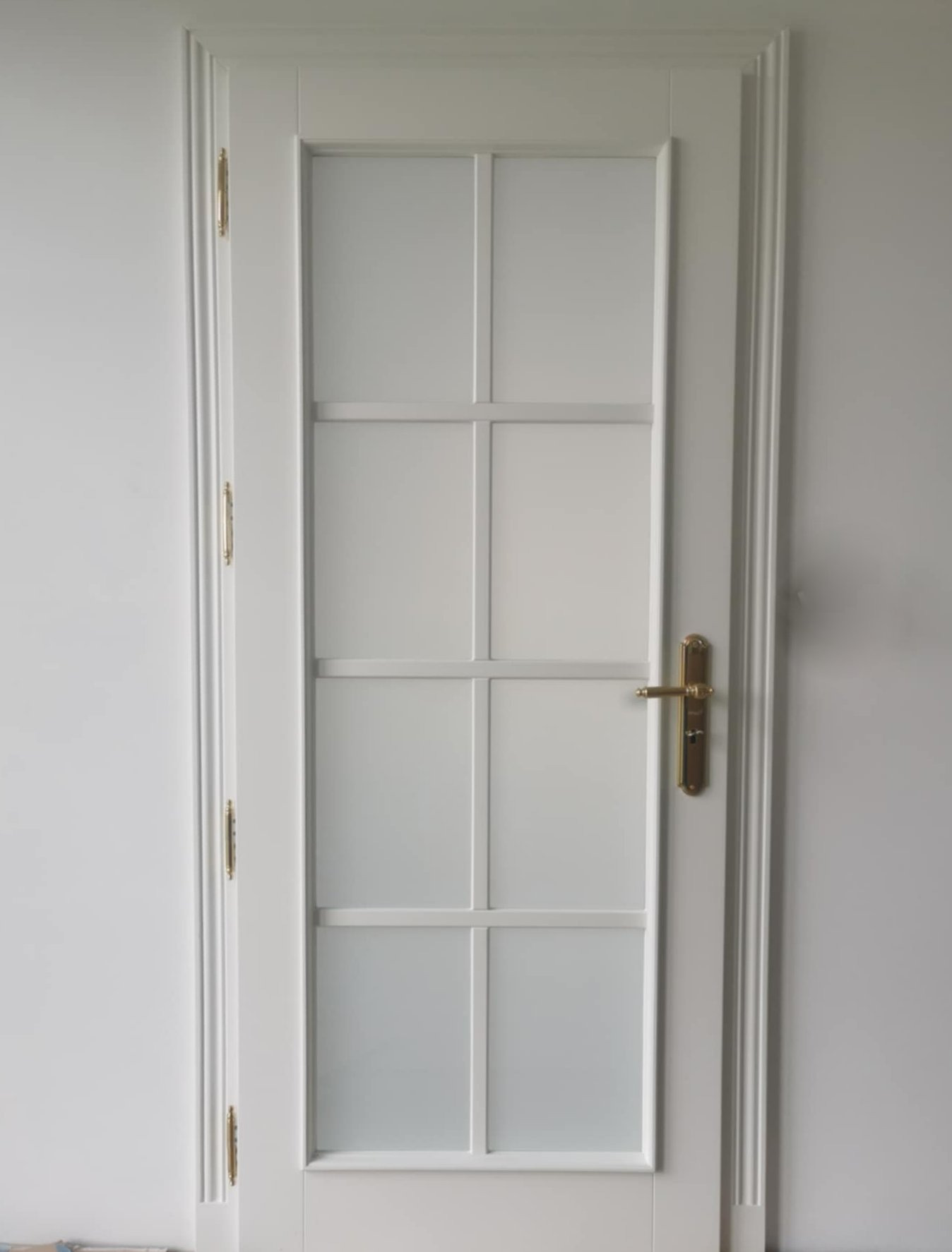 Białe, przeszklone drzwi wewnętrzne z ozdobnymi listwami i złotą klamką, zamontowane w jasnym pomieszczeniu. Ujęcie centralne, prezentujące detale wykończenia.