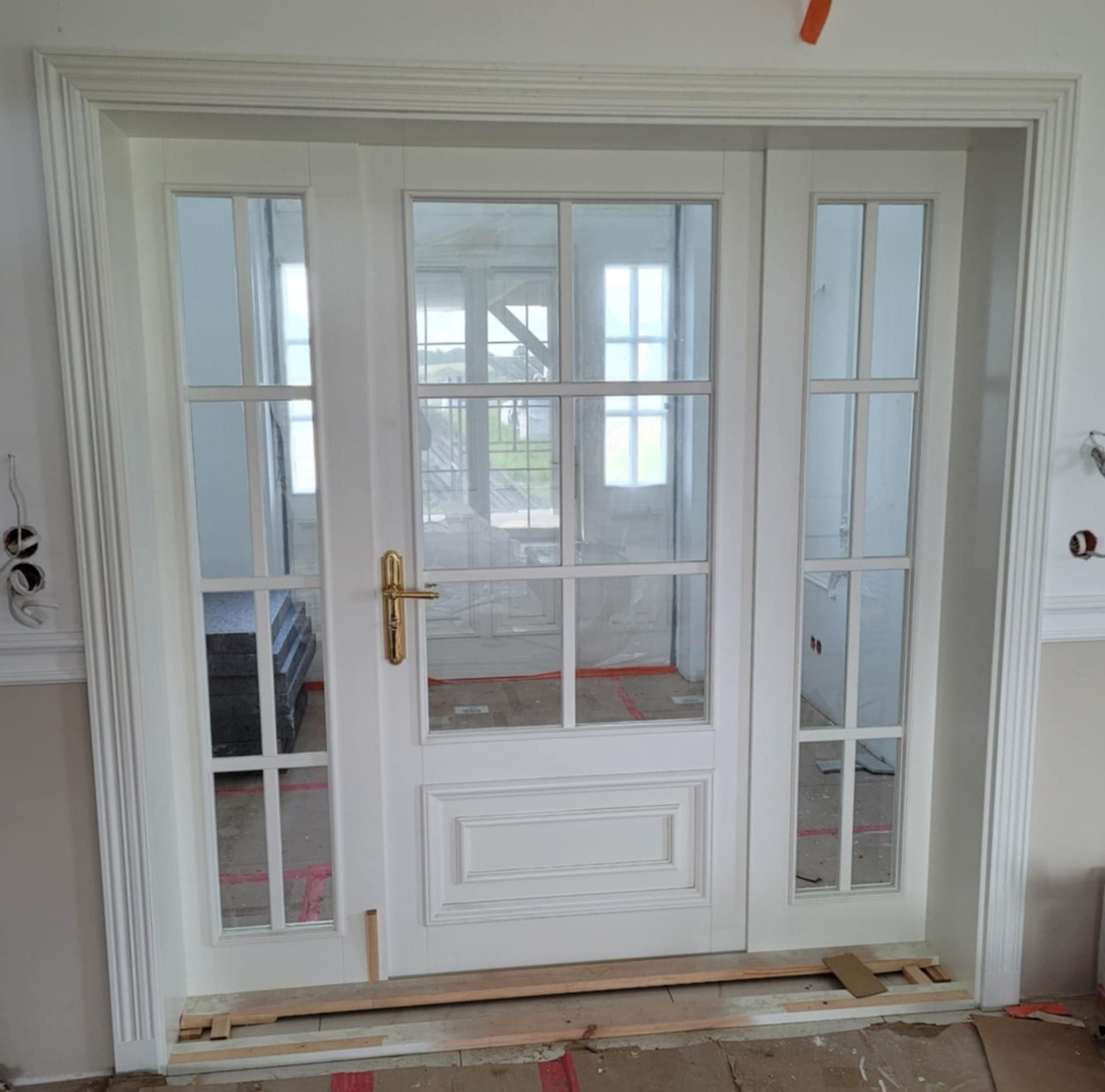 Białe, przeszklone drzwi wewnętrzne z ozdobnymi listwami, zamontowane w surowym wnętrzu. Widoczne przygotowania do dalszych prac wykończeniowych.