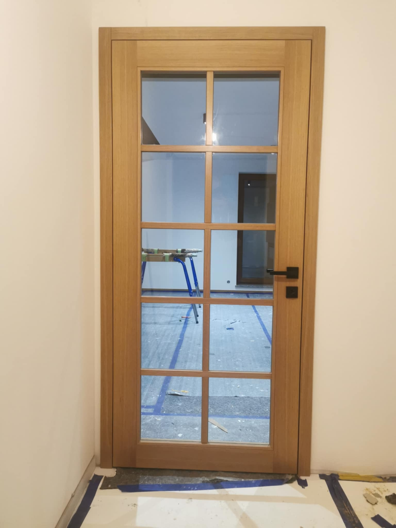 Dębowe drzwi wewnętrzne z szybami w szprosach, czarna klamka, widok na zabezpieczone folią podłoże i kozły malarskie w pomieszczeniu.