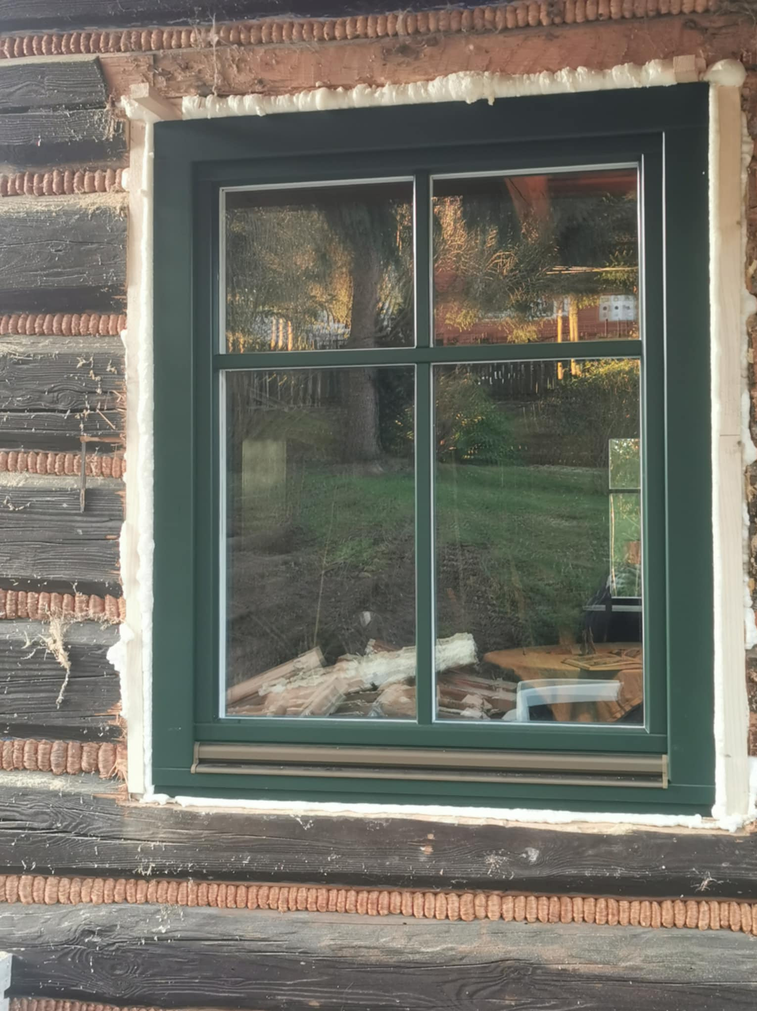 Zamontowane okno z zieloną ramą w drewnianej ścianie domu. Widoczne uszczelnienie pianką montażową. W szybach odbija się zieleń ogrodu.