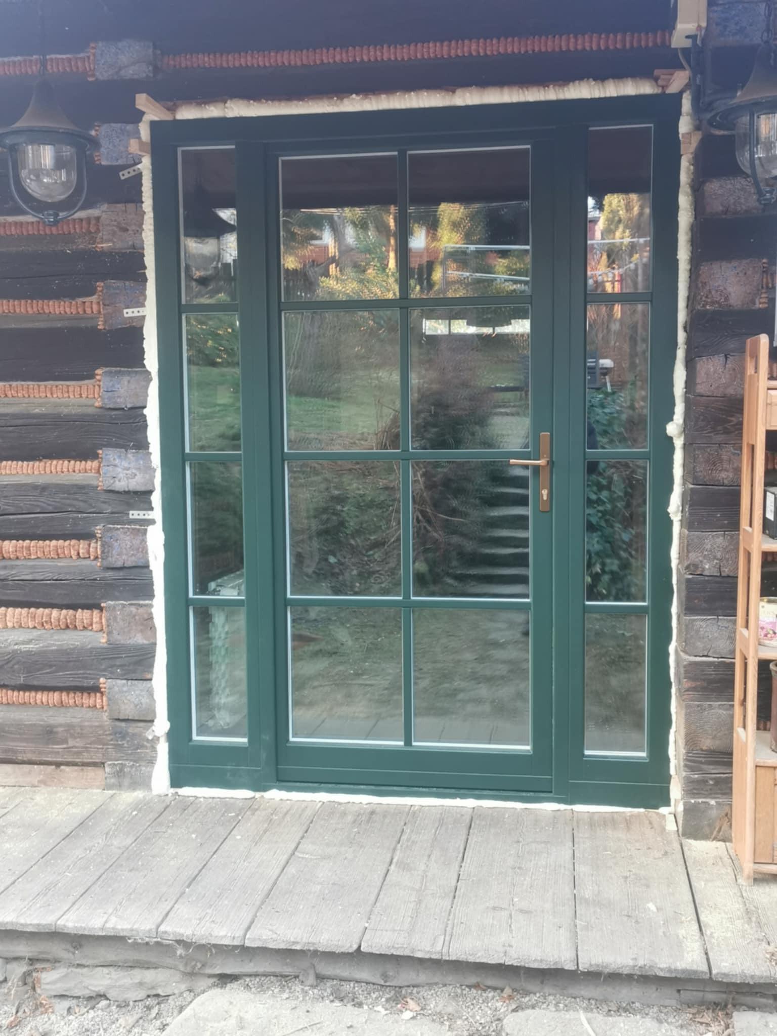 Zielone, przeszklone drzwi zewnętrzne z ozdobnymi szprosami, zamontowane w drewnianym budynku z widoczną pianką montażową.