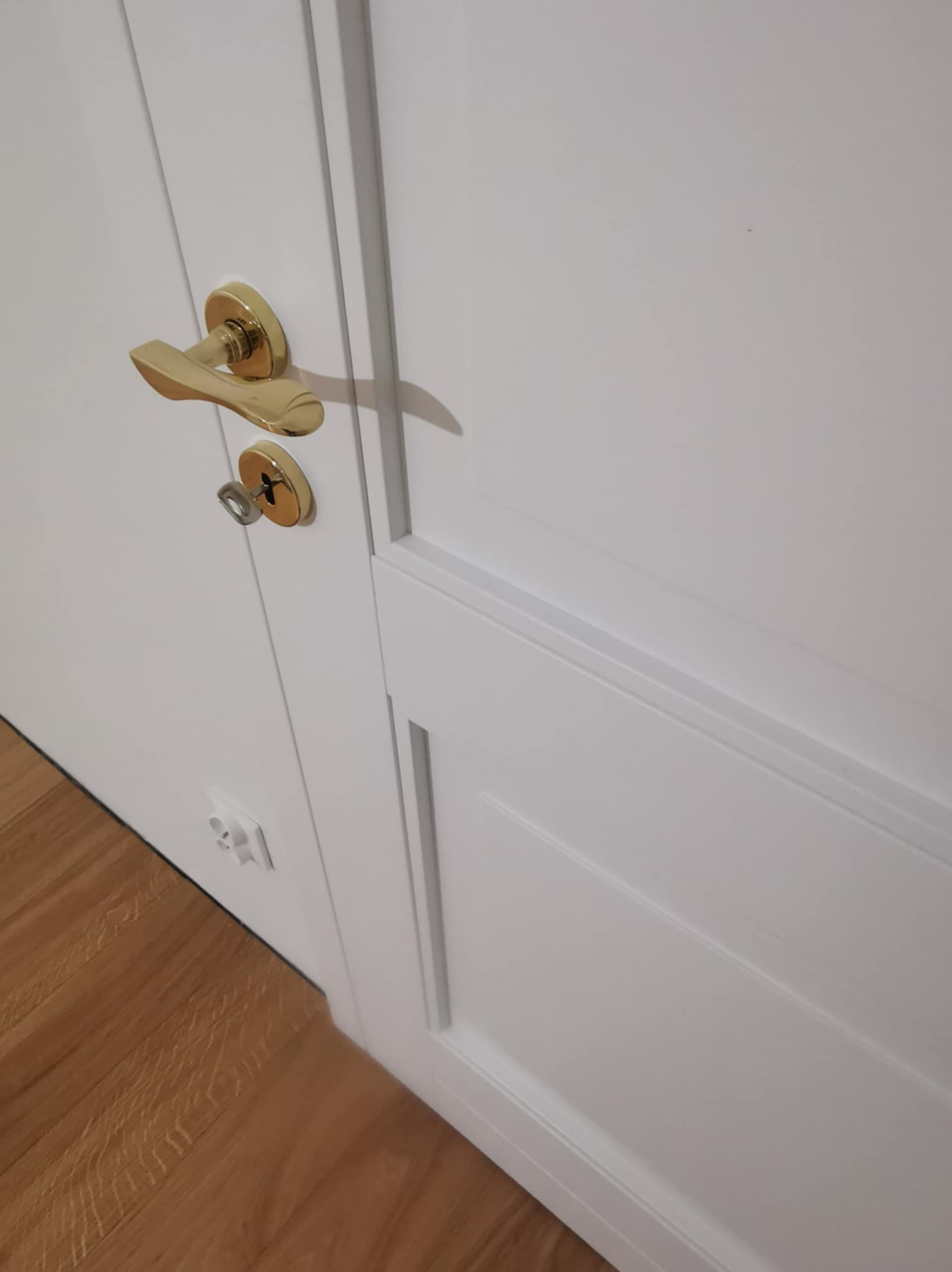 Białe drzwi wewnętrzne z ozdobnymi frezami, złota klamka z kluczem w zamku. Widoczny fragment podłogi z parkietu i gniazdko elektryczne.