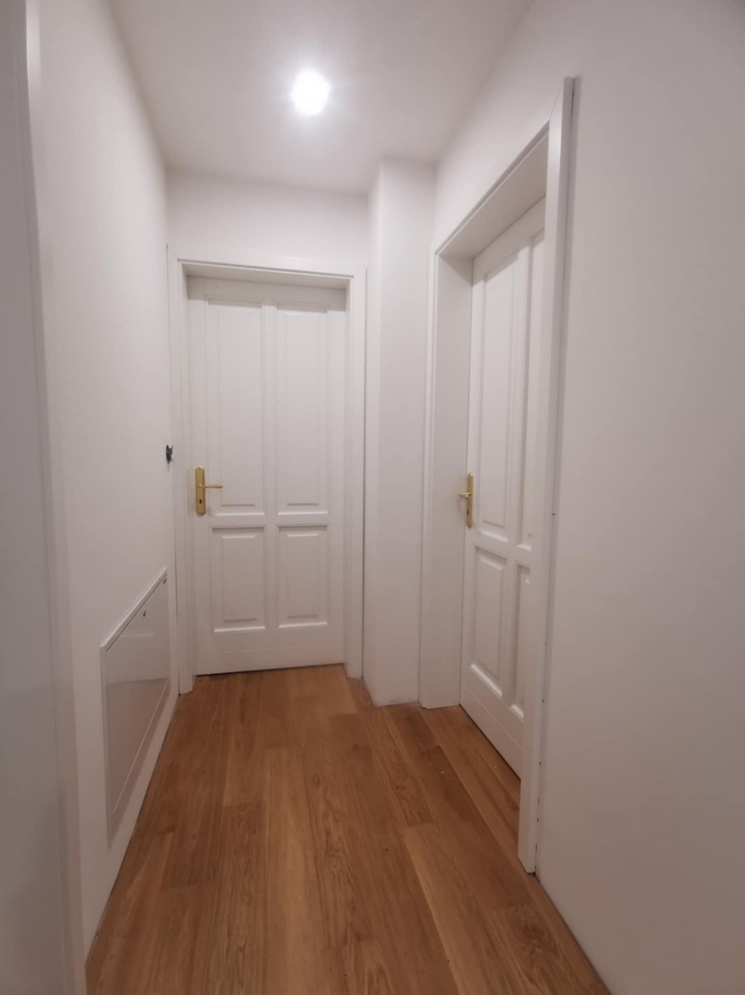 Białe, klasyczne drzwi wewnętrzne z złotymi klamkami w korytarzu z drewnianą podłogą. Jedne drzwi są otwarte, ukazując wnętrze pomieszczenia.