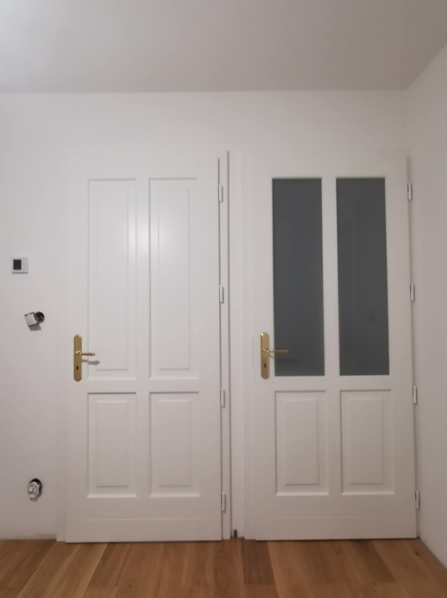 Dwa białe, skrzydłowe drzwi wewnętrzne z złotymi klamkami, jedno z przeszkleniami, na tle białej ściany i drewnianej podłogi. Widoczne gniazdko elektryczne.