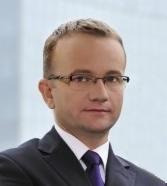 Portret mężczyzny w okularach i ciemnym garniturze na rozmytym tle biurowca.