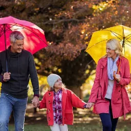Rodzina spaceruje jesiennym parkiem trzymając kolorowe parasole: różowy, żółty. Tata w szarym swetrze i jeansach, mama w czerwonym płaszczu, córka w czerwonej kurtce i szarej czapce.