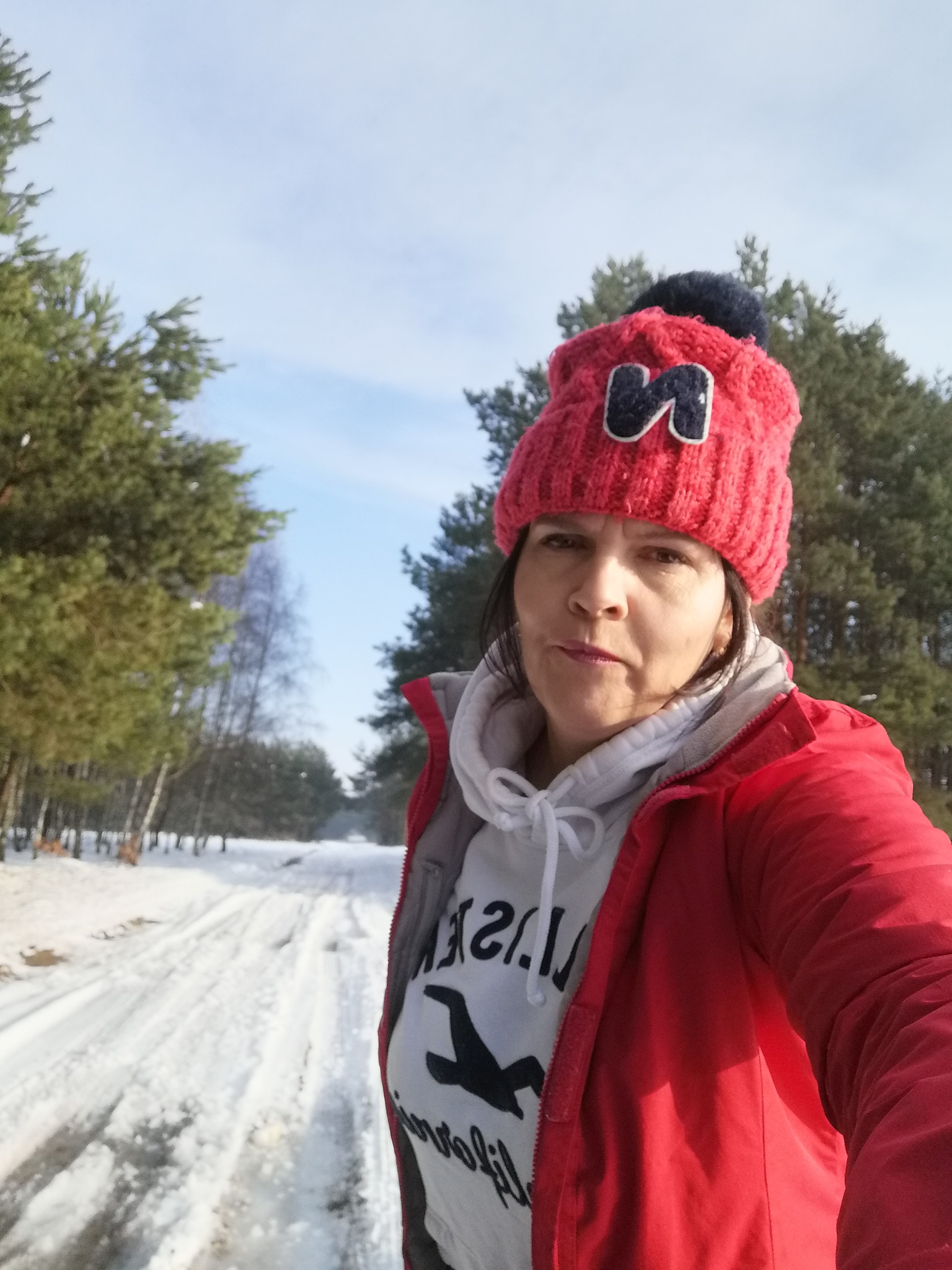 Autoportret kobiety w czerwonej kurtce i czapce z pomponem na tle zimowego krajobrazu z zalesioną drogą pokrytą śniegiem.