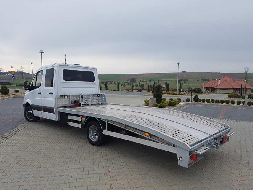 Biały samochód dostawczy z platformą lawety, widok z tyłu, na parkingu z kostki brukowej, w tle zielone wzgórza i budynek z czerwoną dachówką.