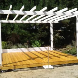 Biała pergola z drewnianym podestem w trakcie budowy, widoczne podpory i czarna folia, tło stanowi zieleń ogrodu.