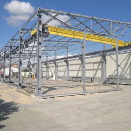 Stalowa konstrukcja hali w trakcie budowy z zamontowaną żółtą suwnicą, widoczne elementy mocujące i jasne niebo w tle.