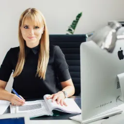 Kobieta w czarnej sukience siedzi przy biurku, pisząc w otwartym notatniku; na biurku dokumenty, komputer iMac i laptop