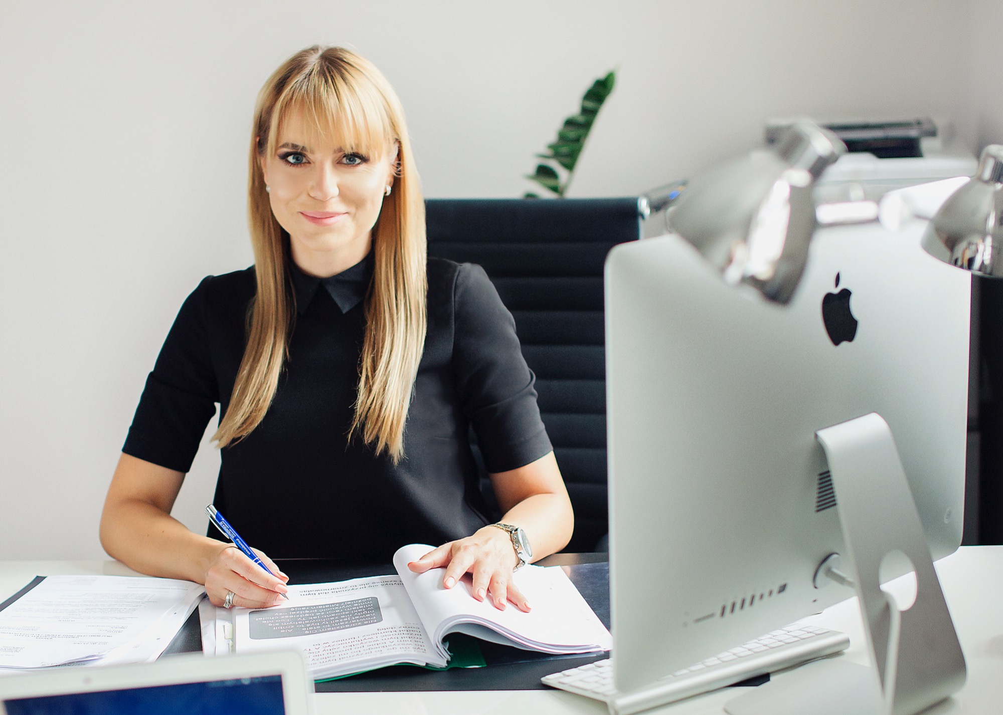 Kobieta w czarnej sukience siedzi przy biurku, pisząc w otwartym notatniku; na biurku dokumenty, komputer iMac i laptop