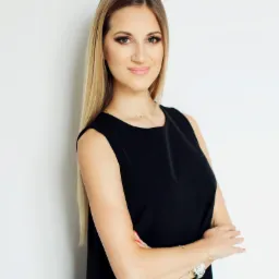 Portret uśmiechniętej blondynki w czarnej sukience, z założonymi rękami, na białym tle, z delikatnym makijażem i biżuterią.