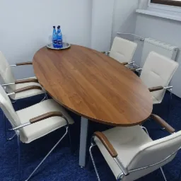 Sala konferencyjna z owalnym drewnianym stołem, sześcioma krzesłami z kremowej skóry i chromowanymi nogami, oraz butelkami wody mineralnej na srebrnej tacy.