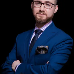 Portret mężczyzny w okularach, niebieskim garniturze i koszuli w kratę, z ozdobną poszetką w kieszonce, na czarnym tle.