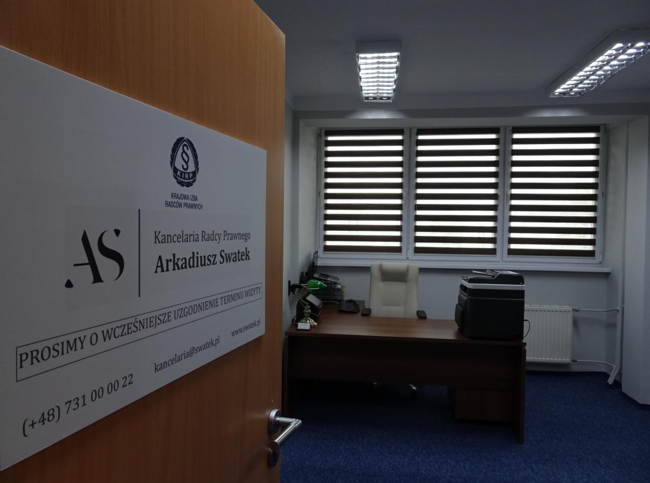 Tabliczka informacyjna na drzwiach do kancelarii radcy prawnego Arkadiusza Swatka, widok na biuro z biurkiem, krzesłem i drukarką w tle.