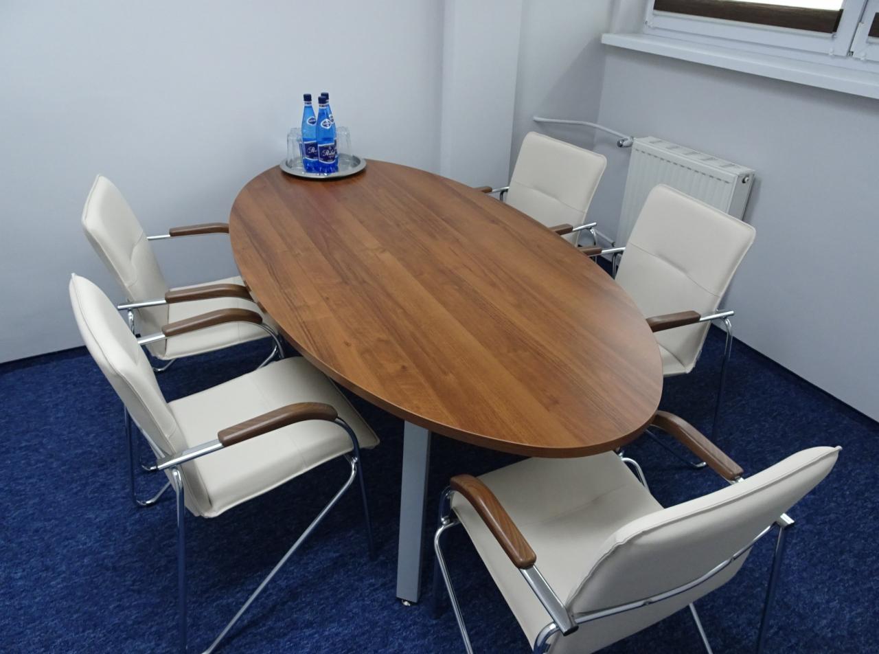 Sala konferencyjna z owalnym drewnianym stołem, sześcioma krzesłami z kremowej skóry i chromowanymi nogami, oraz butelkami wody mineralnej na srebrnej tacy.