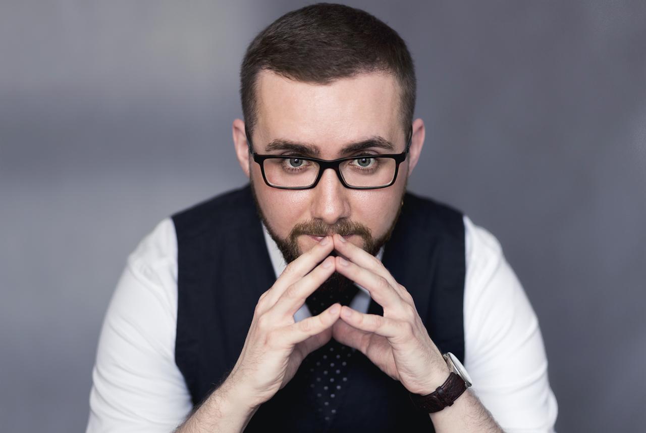 Portret mężczyzny w okularach, z zamyślonym spojrzeniem i splecionymi dłońmi przed twarzą, ubrany w białą koszulę i ciemną kamizelkę.