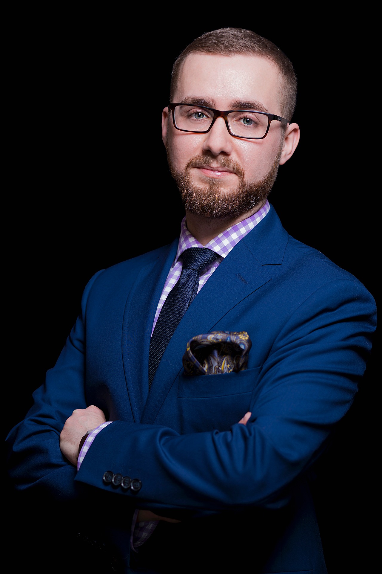 Portret mężczyzny w okularach, niebieskim garniturze i koszuli w kratę, z ozdobną poszetką w kieszonce, na czarnym tle.