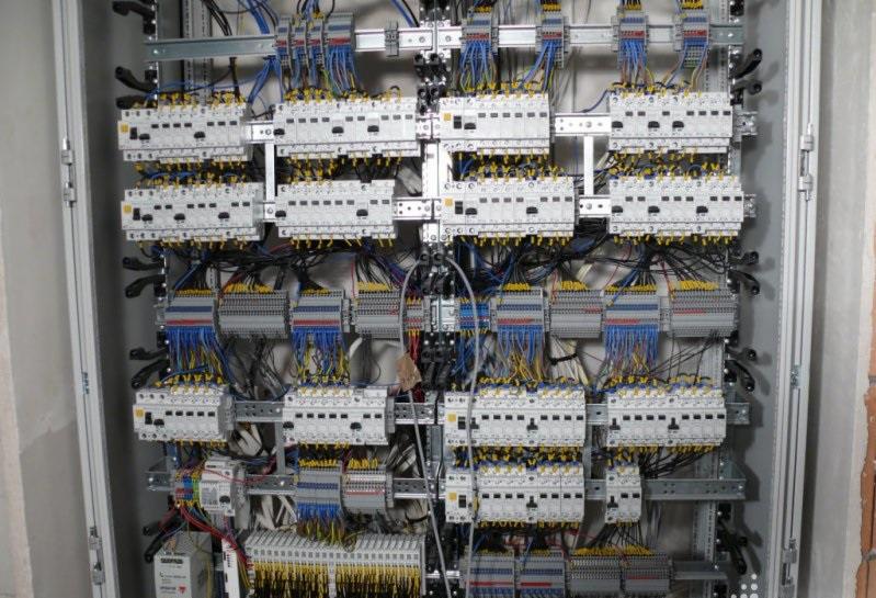 Rozbudowana szafa elektryczna z licznymi modułowymi wyłącznikami i zabezpieczeniami, okablowanie w kolorowych izolacjach, zamontowana w surowym pomieszczeniu z widocznym fragmentem ściany.