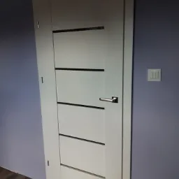 Białe drzwi wewnętrzne z czterema poziomymi czarnymi wstawkami, zamontowane w ścianie o fioletowym odcieniu, widoczny fragment podłogi z paneli.