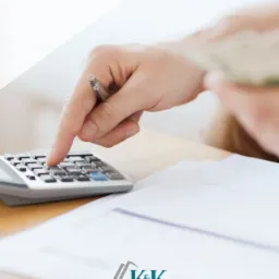 Osoba liczy na kalkulatorze, trzymając w drugiej ręce plik banknotów, na biurku leżą dokumenty.